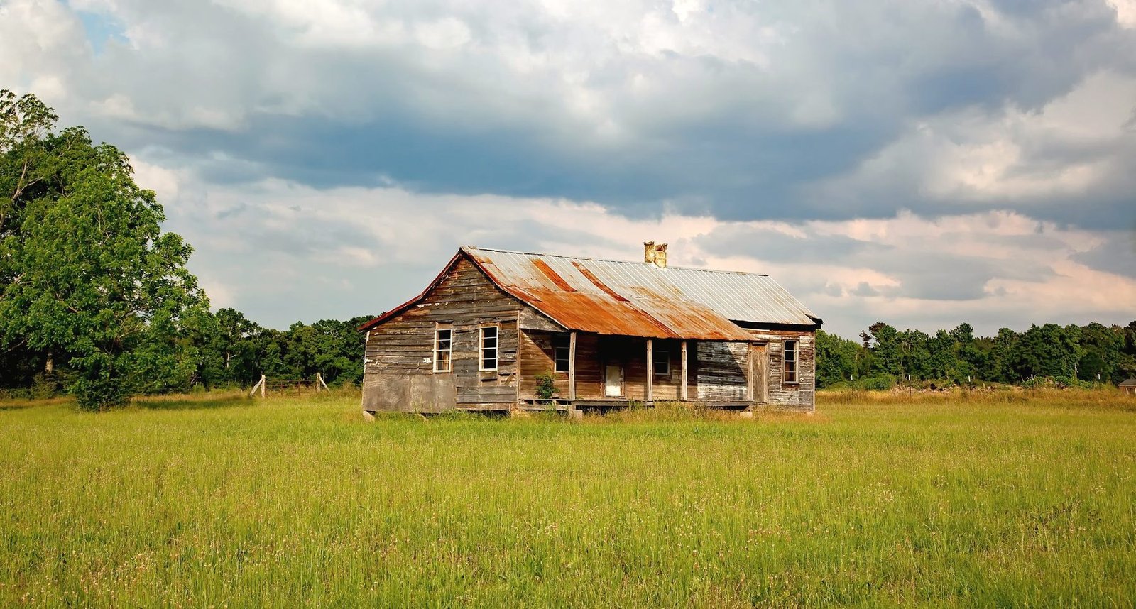 An old wooden farmhouse with a rusted tin roof standing alone in a grass field.